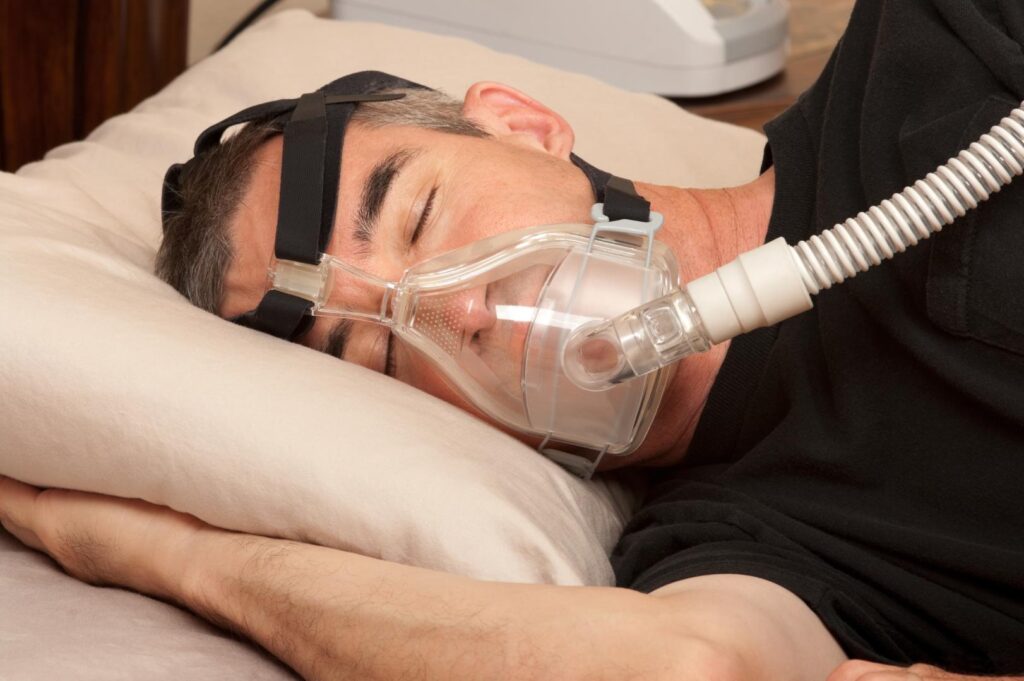 Man sleeps on his side wearing a clear full-face CPAP mask connected to a flexible air hose.